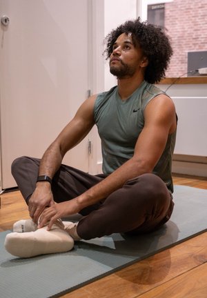 Hombre con camiseta sin mangas y pantalones deportivos sentado con las piernas cruzadas sobre una esterilla de yoga, estirando los pies en el interior sobre un suelo de madera.