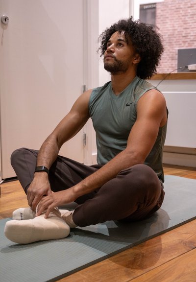 Hombre con camiseta sin mangas y pantalones deportivos sentado con las piernas cruzadas sobre una esterilla de yoga, estirando los pies en el interior sobre un suelo de madera.
