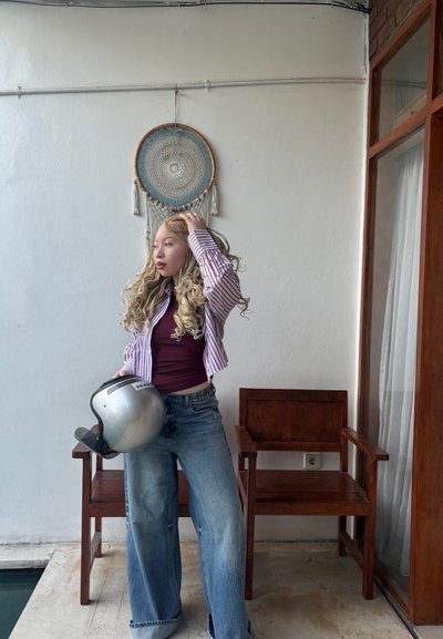 Una persona con una camisa a rayas y corta sobre una blusa granate sostiene un casco plateado, vistiendo jeans anchos azules, con sillas de madera al fondo.