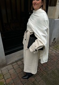 Šviesiai ruda tranšėja su juodais mygtukais, dėvima ant baltos šalikos; juoda rankinė laikoma po ranka; smaili juoda avalynė; akmeninė danga.