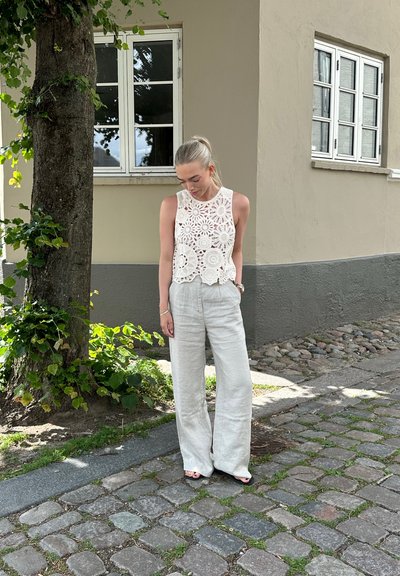 Top de crochet blanco con cortes en forma de flor, combinado con pantalones de lino gris claro de pierna ancha. El entorno presenta una superficie de piedra y vegetación.