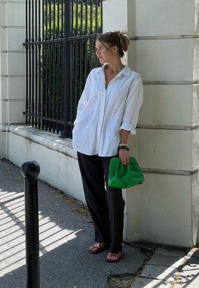 Camisa de lino blanca oversize, pantalones negros a rayas, sandalias rojas y un bolso verde con pliegues, posando cerca de una pared blanca y una barandilla negra.
