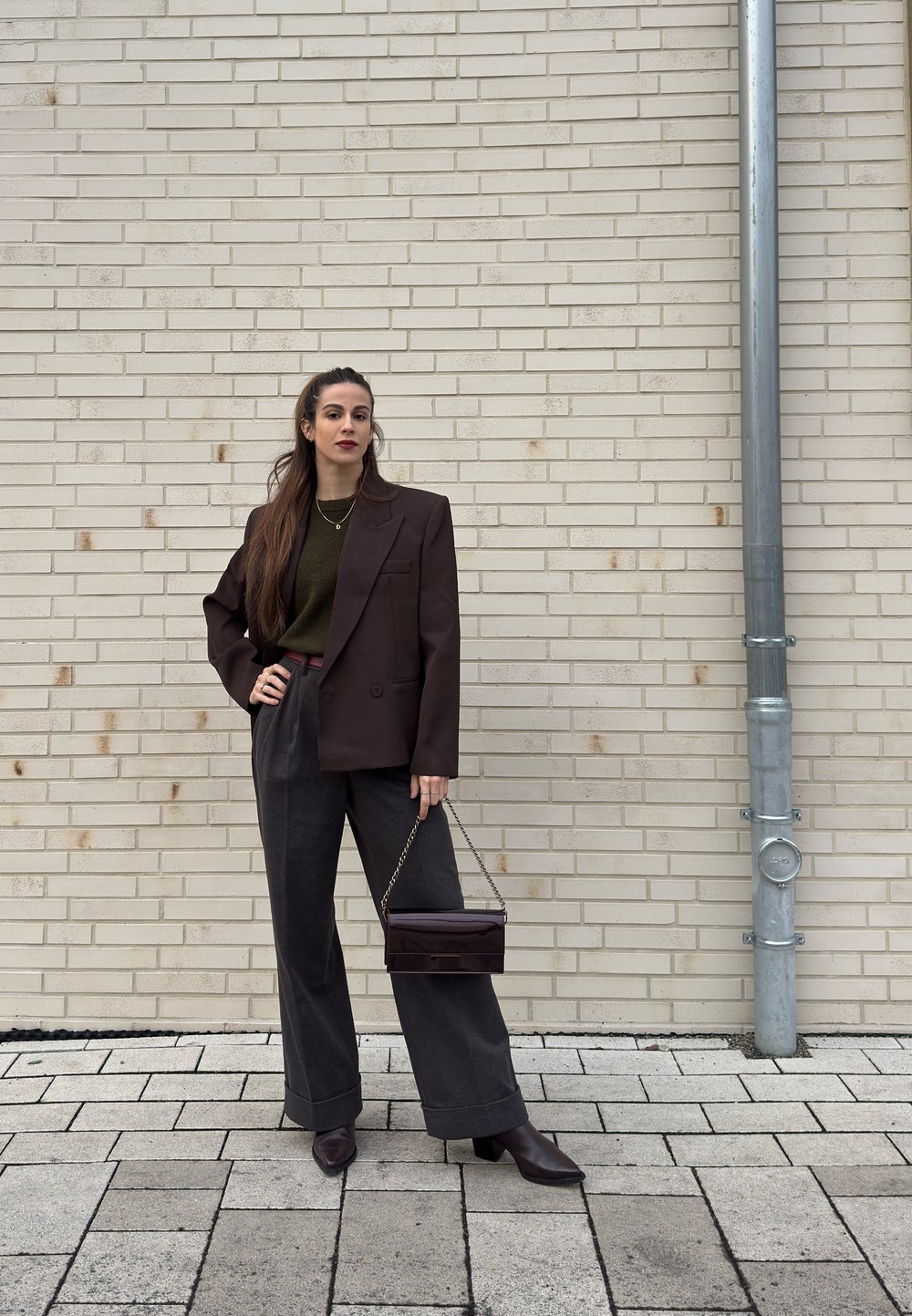 Femme en blazer foncé et pantalon à jambes larges tenant un petit sac à chaîne, debout contre un mur de briques claires avec un tuyau en métal sur la droite.
