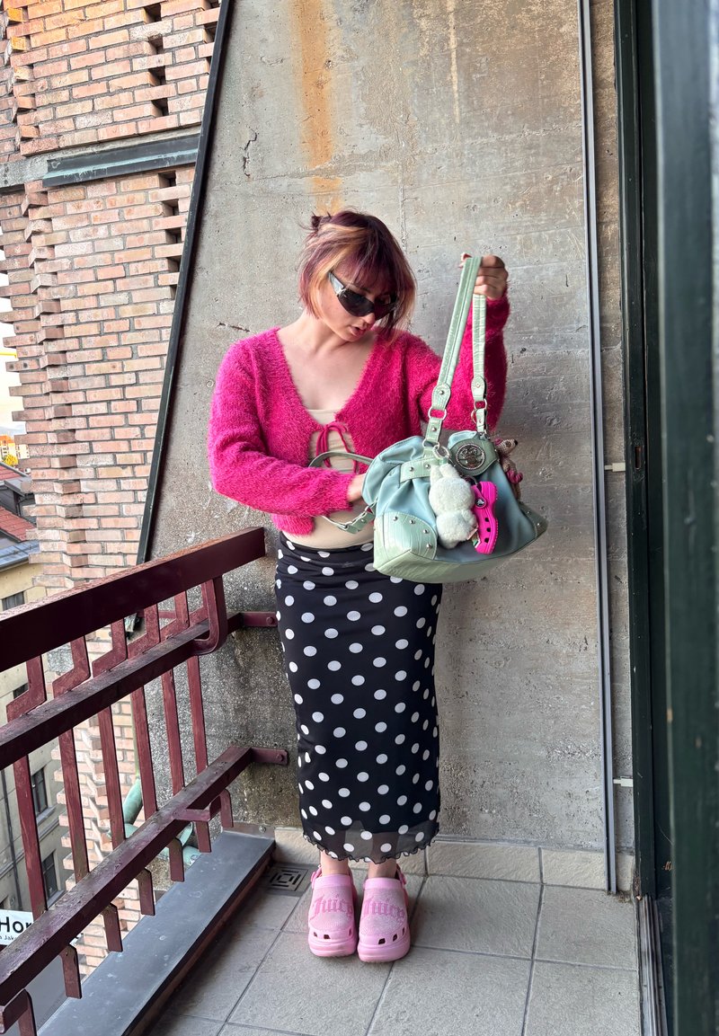 Femme aux cheveux courts, pull rose, jupe à pois et sabots roses, debout sur un balcon tenant un sac à main vert clair avec des accessoires.