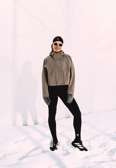 Mujer de pie sobre la nieve, vestida con chaqueta marrón, mallas negras, diadema, guantes, gafas de sol y zapatillas de correr negras, sonriendo a la cámara.