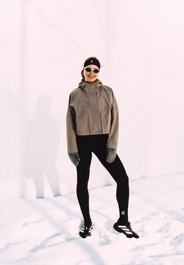 Femme debout sur la neige portant une veste marron, des leggings noirs, un bandeau, des gants, des lunettes de soleil et des chaussures de course noires, souriant à la caméra.
