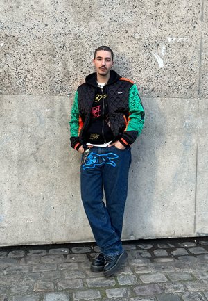 Chaqueta negra acolchada con mangas verdes y naranjas, superpuesta a una sudadera oscura. Gráfico azul en unos vaqueros oscuros, zapatos negros, de pie contra una pared de hormigón.