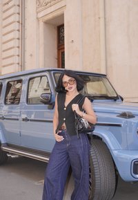 Haut sans manches noir avec des boutons dorés, pantalon large en denim bleu, et un sac à main noir. SUV bleu clair garé en arrière-plan.