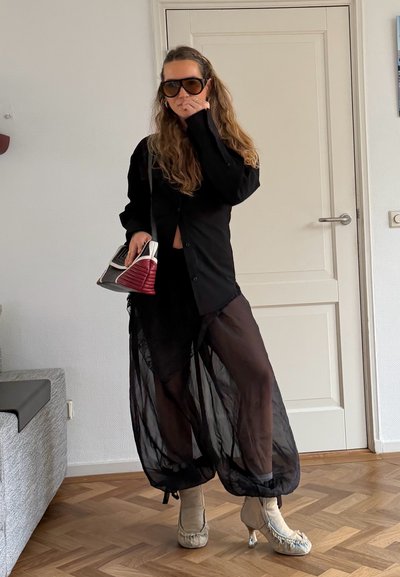 Mujer con camisa negra de gran tamaño, pantalones anchos negros transparentes, tacones beige, gafas de sol grandes, sujetando un bolso rojo y blanco en interiores.