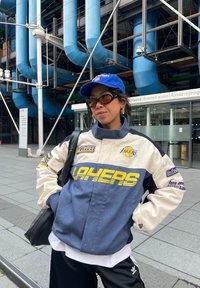 Veste Lakers en bleu marine et crème, avec un logo jaune, une coupe oversize et des patchs en caoutchouc. Le modèle porte des lunettes de soleil et une casquette bleue.
