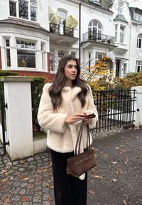Jeune femme en manteau de fourrure crème tenant un sac à main marron et des lunettes de soleil dans une rue pavée avec des maisons blanches et des feuilles d'automne en arrière-plan.