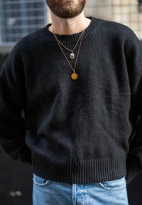Suéter negro de punto con dobladillos y puños acanalados, combinado con tres collares dorados y de colores, acompañado de vaqueros de color azul claro.