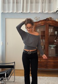 Grijs off-shoulder cropped topje met lange mouwen, gecombineerd met zwarte flared jeans en een zilverkleurige riem met gesp, geplaatst in een neutrale interieuromgeving.