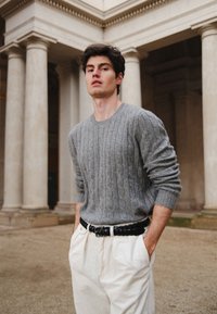 Pull gris à torsades en tissu texturé, associé à un pantalon en velours côtelé blanc et une ceinture noire tressée. En arrière-plan, des colonnes classiques.