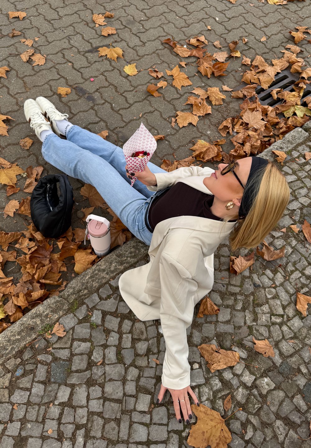 Witte blazer over een donker shirt, lichtblauwe spijkerbroek, witte sneakers; houdt een roze-witte papieren kegel met snacks vast. Kasseien achtergrond met herfstbladeren.