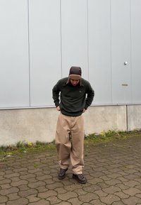 Khaki bő fazonú nadrág, sötétzöld pulóver és barna prémvadász sapka. A ruha laza szabású és texturált kötött mintával rendelkezik. Barna túracipő.