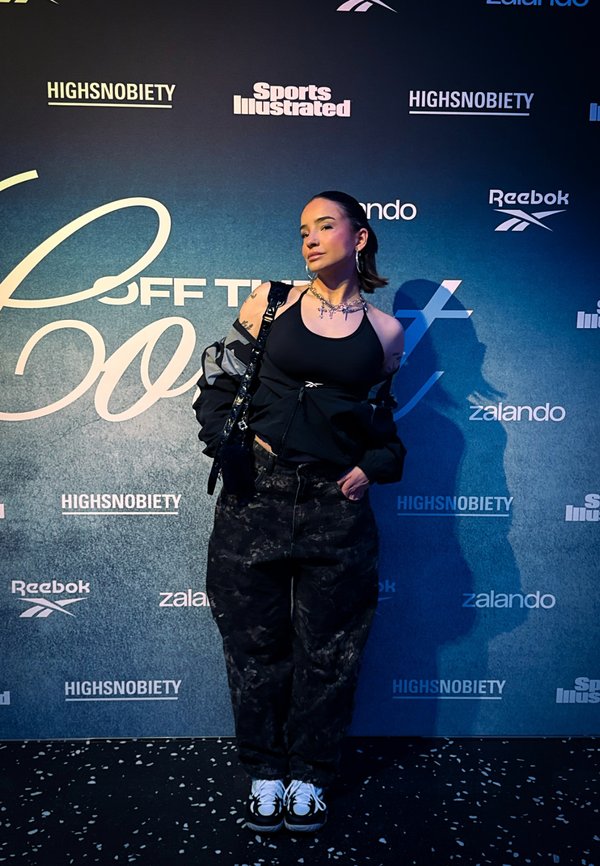 Jeune femme posant en débardeur noir, pantalon camouflage et baskets devant un fond d'événement avec des logos, notamment Reebok et Sports Illustrated.