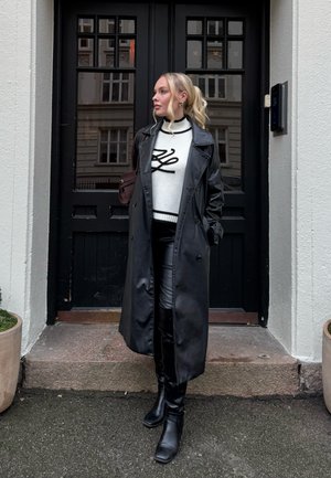 Mujer con cabello rubio recogido en una cola de caballo, lleva un abrigo largo de cuero negro, suéter blanco con diseño negro, pantalones negros y botas, de pie junto a una puerta negra.