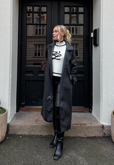 Mujer con cabello rubio recogido en una cola de caballo, lleva un abrigo largo de cuero negro, suéter blanco con diseño negro, pantalones negros y botas, de pie junto a una puerta negra.