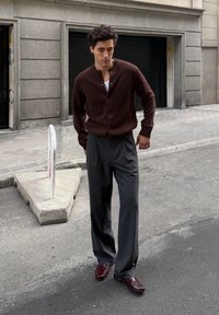 Bruine gebreide cardigan, grijze krijtstreep broek, donker bordeauxkleurige schoenen; staand op straat nabij een grijze stenen gebouw en het trottoir.