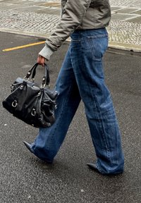Blaue weit geschnittene Jeans, graue Bomberjacke mit gerippten Bündchen, schwarze Lederhandtasche mit silbernem Beschlag und spitze schwarze Schuhe.