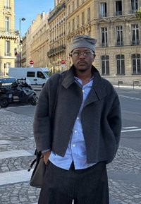 Szürke gyapjú dzseki, repesztett gallérral, kék csíkos ing rétegezve alatta, sötét nadrággal párosítva. Laza napszemüveg és szürke fejkendő.