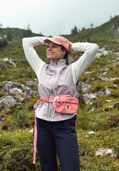 Chaleco de cierre en color lavanda claro con bolsillos, camisa blanca de manga larga, pantalones negros y una riñonera rosa con un logo. Fondo de paisaje verde.