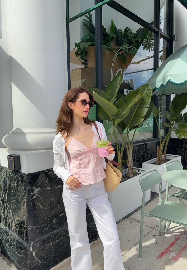 Mujer con gafas de sol sostiene un batido verde, lleva top rosa, pantalones blancos y cárdigan, de pie cerca de una cafetería al aire libre con plantas y sillas.
