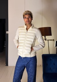 Άτομο με ξανθά σγουρά μαλλιά που φοράει κρεμ καπιτονέ μπουφάν και μπλε παντελόνι με σχέδια, στέκεται μέσα στο σπίτι κοντά σε μια λάμπα και έναν σκουρομπλε καναπέ.