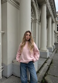Maglione oversized rosa chiaro con polsini a coste, abbinato a jeans in denim blu a gamba larga. Sullo sfondo si vedono colonne classiche e pavimentazione grigia.