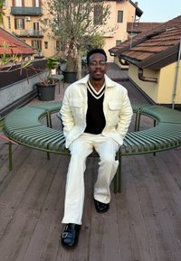 Persona seduta su una panchina curva in metallo verde. Indossa una giacca bianca texture sopra un maglione nero a scollo a V e pantaloni bianchi; scarpe nere.