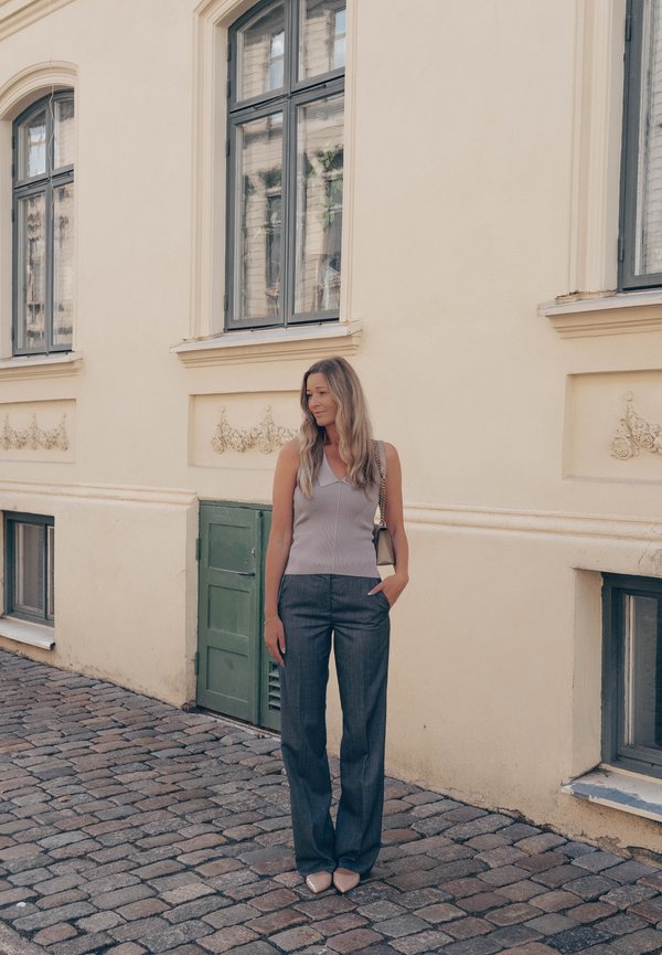 Mujer con el pelo largo, vestida con una camiseta sin mangas y pantalones grises, está de pie en una calle empedrada junto a un edificio beige con grandes ventanas.