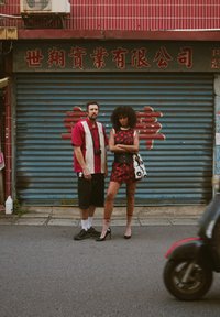 Un hombre con una camisa roja y blanca que sostiene una cámara está al lado de una mujer con un vestido floral rojo y tacones negros, ambos frente a un fondo de persiana azul.