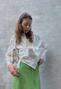 Chemise blanche à manches longues avec broderies florales, superposée à un haut blanc transparent, associée à une jupe verte avec un détail de cordon.