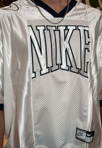 Bílé Nike tričko s prodyšnou tkaninou, velkým černým logem "NIKE", námořnickými akcenty, výstřihem do V a ochrannou známkou na dolní části přední strany.