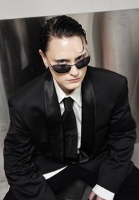Smoking noir avec revers en satin, chemise blanche et cravate. Lunettes de soleil foncées, cheveux coiffés en arrière et boucle d'oreille discrète. Fond argenté.
