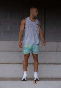 Grå atletisk tanktopp med bølgete mønster; lyse aqua shorts med geometriske former; hvite joggesko; står på betongtrapper.