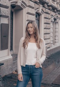 Jasnobeżowy kardigan na biały top, zestawiony z niebieskimi dżinsami. Miękka faktura, dopasowany krój i ściągacze przy rękawach. Miejska sceneria, tło ze ścianą z kamienia.