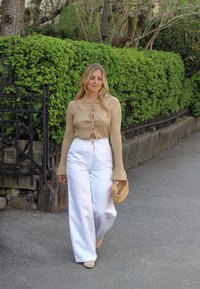 Beige geribbelde langemouwen top met een kraag en een detail van een strik aan de voorkant; witte wijde broek; houdt een gevlochten clutch vast; groene bladeren op de achtergrond.