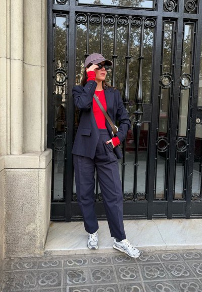 Mujer con blazer y pantalones azul marino, camisa roja, gorra gris, gafas de sol y zapatillas blancas, de pie frente a una verja de metal negro ornamentada.