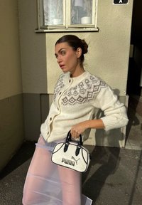 Cardigan crema a maglia grossa con giogo decorato, abbinato a una borsa nera e bianca con logo Puma e gonna rosa trasparente.
