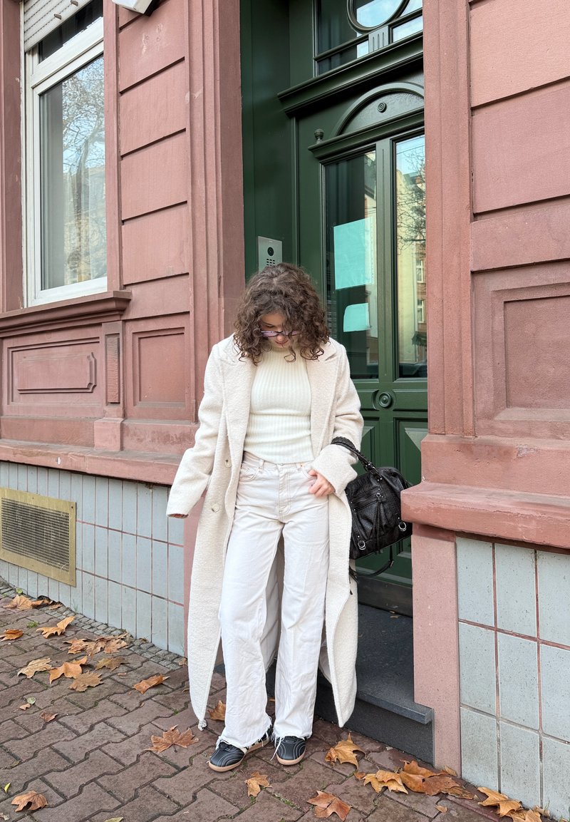 Manteau crème texturé porté sur un pull léger et un pantalon pâle. Sac à main noir et coiffure stylée. Debout près d'une porte verte avec un mur en briques.