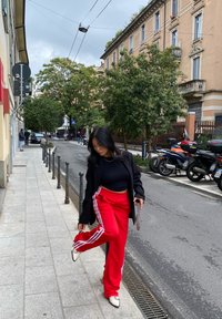 Röda träningsbyxor med vita ränder, svart crop top och en svart kavaj. Hon står på en trottoar nära parkerade motorcyklar. Molnig himmel.