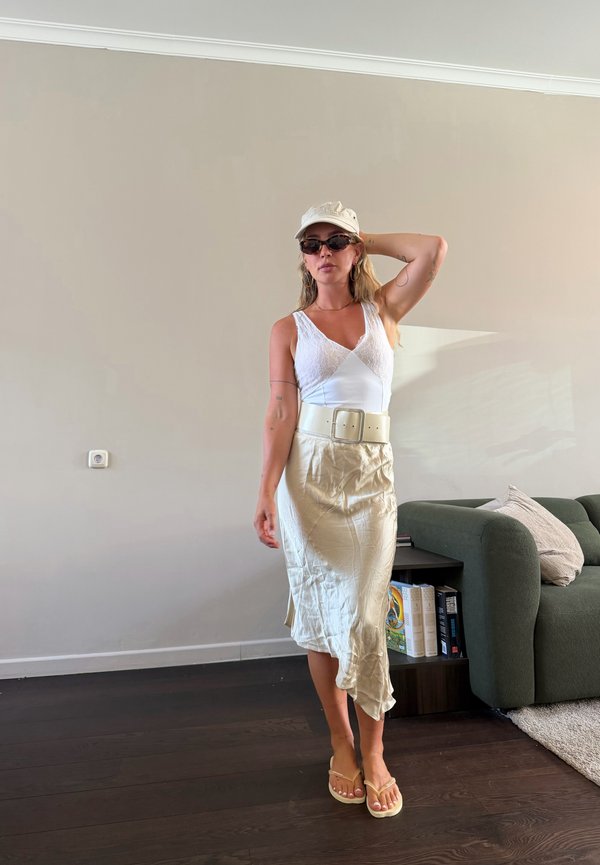 Femme portant un haut en dentelle blanche, une jupe beige, des lunettes de soleil et une casquette, debout à l'intérieur près d'un canapé vert avec des livres sur une table d'appoint.