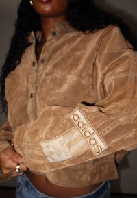 Brązowa zamszowa kurtka z zapinanym na guziki kołnierzem, trzema paskami wzdłuż rękawów oraz wyraźną metką Adidas na mankiecie.