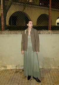 Bronzová vlnená bunda cez svetlozelené vrstvené šaty, doplnená čiernymi členkovými čižmami. Stojí pri nízkej stene s pozadím z pletiva.