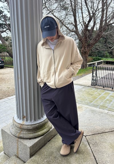 Sudadera beige con cremallera, gorra azul marino con el texto "DOG DAD", pantalones oscuros y zuecos color beige claro, apoyado contra una columna cerca de un camino de piedra.