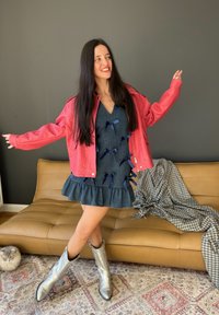 Chaqueta de cuero sintético roja, vestido de mezclilla con lazos azul marino, dobladillo con volante y botas de vaquero plateadas. Con fondo de una pared oscura y un sofá beige.