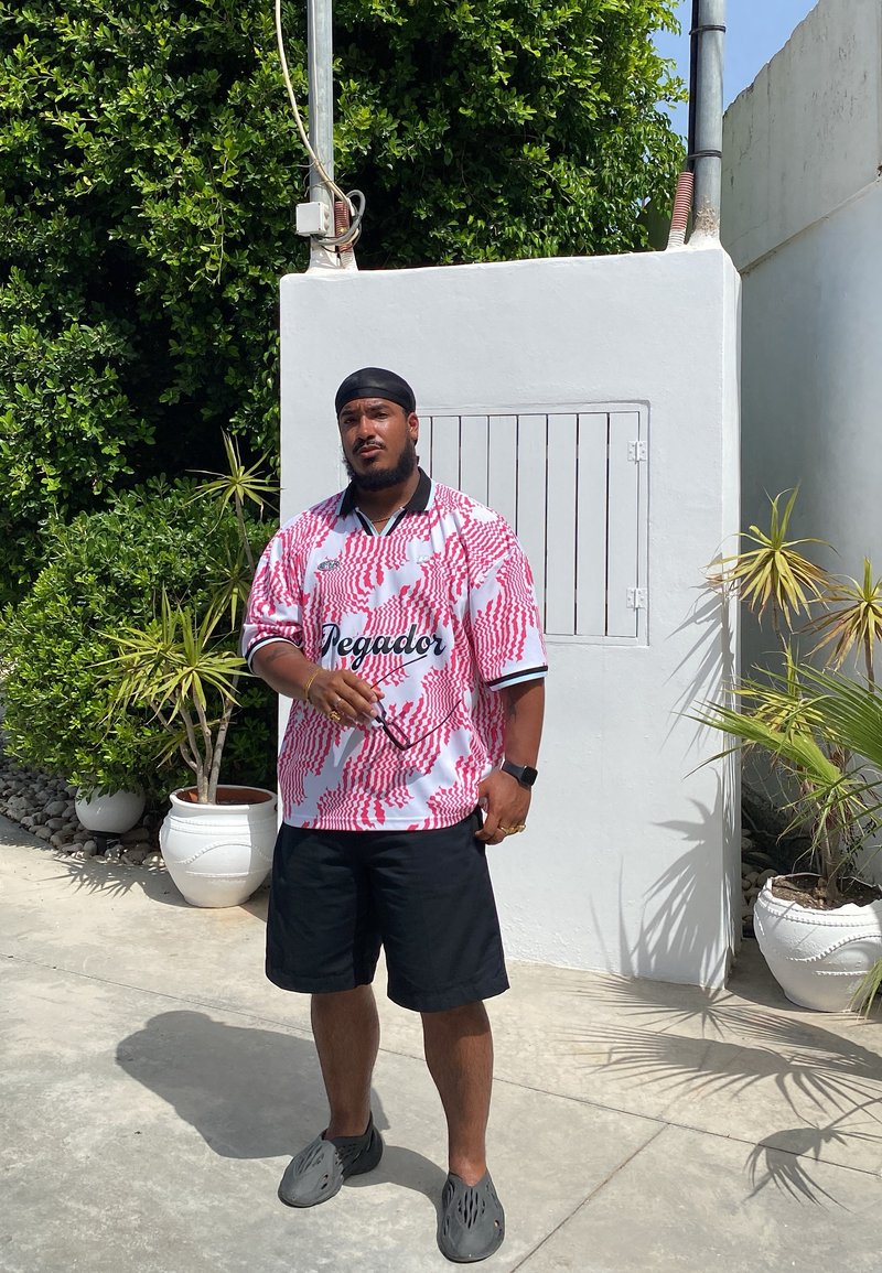 Κόκκινο και λευκό πουκάμισο με μοτίβο και το κείμενο "Teguidor", μαύρη βερμούδα και γκρι παντόφλες. Στηρίζει τον εαυτό του σε έναν λευκό τοίχο και στο πράσινο.