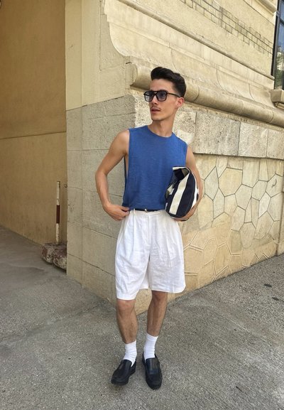 Hombre con camisa sin mangas azul, pantalones cortos blancos, gafas negras, sosteniendo una bolsa de rayas, de pie junto a una pared de piedra en la acera.